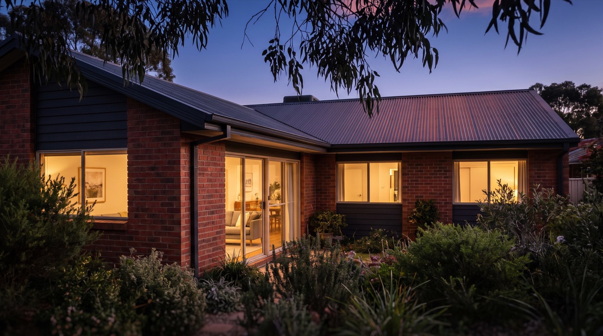 A brick home with a Colorbond roof and lit windows at dusk behind a planted garden