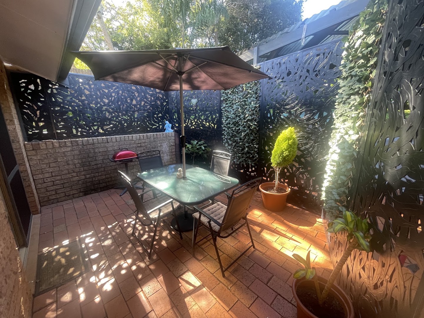 A paved courtyard with an outdoor table, umbrella and potted plants in late sun