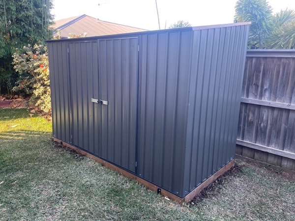 A steel garden shed standing closed on a level base beside a timber fence